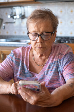 An elderly woman wearing glasses sits at a kitchen table and concentrates on her smartphone, scrolling through messages. The senior appears focused and calm, illustrating modern technology use, 