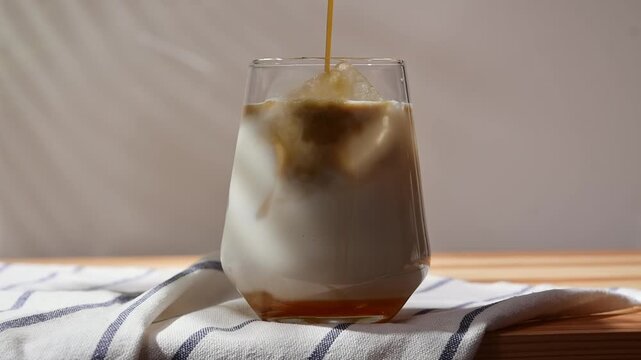Hojicha pours into oat milk over ice in clear glass, creating layered iced latte on wooden table against soft background.