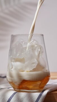 Oat milk pours over ice and golden syrup in clear glass during preparation of chilled homemade hojicha latte on wooden table.
