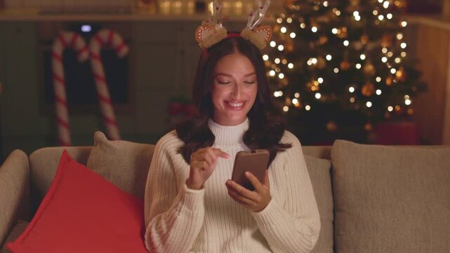 Smiling young woman wearing festive reindeer antlers using smartphone during home Christmas