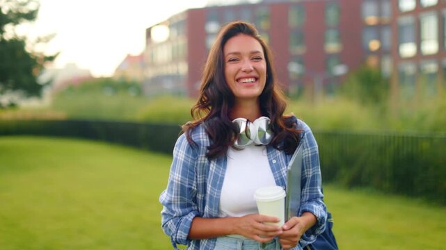 Happy female college student smiling at camera while holding takeaway coffee and laptop, standing outdoors, enjoying campus life. 4K, copy space