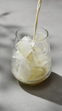 Oat milk pours over ice in glass to prepare cold hojicha latte, showing simple plant based beverage preparation in sunlight.