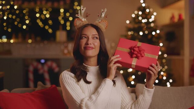 Young woman wearing reindeer antlers holding wrapped Christmas gift, guessing whats inside