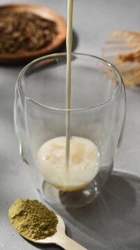 Oat milk is poured into a glass with syrup beside hojicha powder to make a creamy roasted tea latte on a gray stone tabletop.