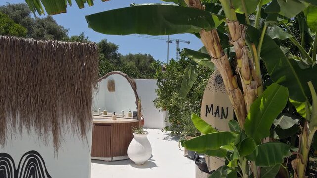 A small arched wall niche stands beside a thatched roof structure surrounded by lush green tropical plants