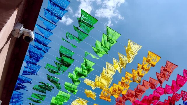 Colorful papel picado decorations in blue, green, yellow, orange, and red hanging in rows against clear blue sky, creating festive Mexican cultural decoration.