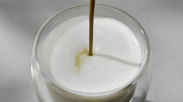 Hojicha concentrate pours into foamed milk in clear glass, creating roasted tea latte pattern in horizontal macro closeup.