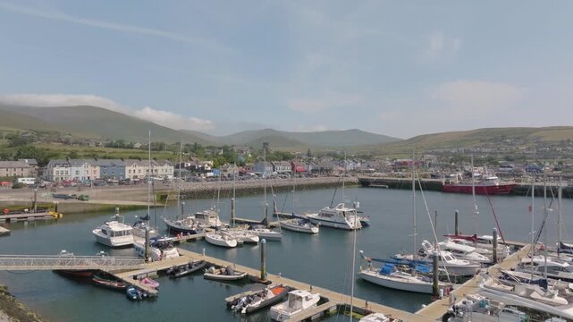 Sailboats and yachts sit moored in Dingle Marina with colorful waterfront buildings and hills beyond on a clear summer day, drone flyover shot.