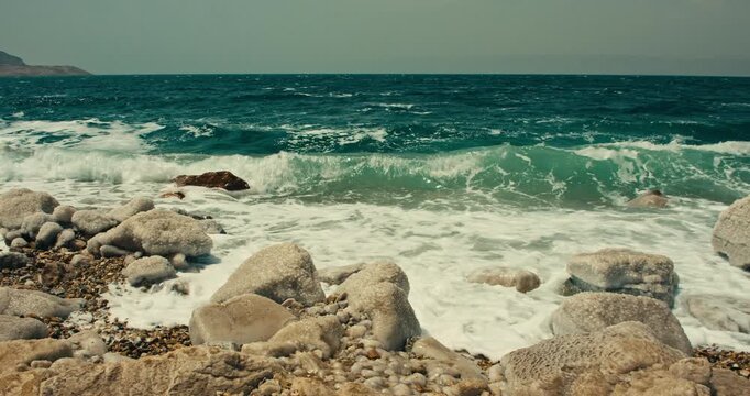 Wide view of Dead Sea waves washing over rocky salt formations in Jordan with
turquoise water, white foam, and a rugged hypersaline shoreline in warm daylight.
