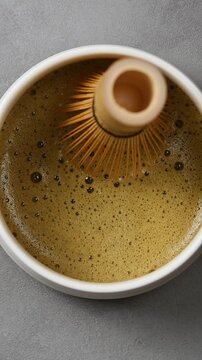 Foamy hojicha tea texture is shown in ceramic bowl as bamboo whisk moves across surface during vertical closeup preparation.