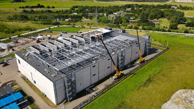 Aerial view of a large new data center facility under construction, showing structural steel frameworks and rooftop cooling systems, with cranes and heavy equipment working on site