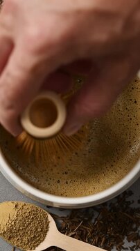 Bamboo whisk moves across foamy hojicha in ceramic bowl, with powder and roasted leaves nearby in vertical macro closeup.