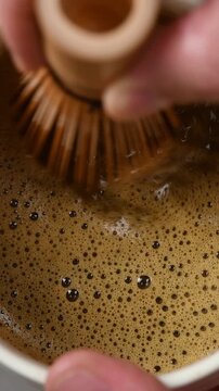 Hand slowly moves bamboo whisk through foamy hojicha in ceramic bowl, showing smooth texture during Japanese tea preparation.
