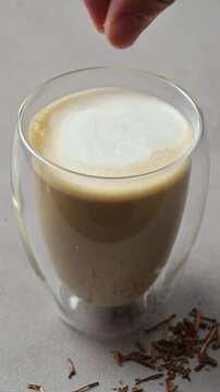 Hand sprinkles sea salt over foamy hojicha oat milk latte in glass, finishing creamy Japanese roasted tea drink preparation.