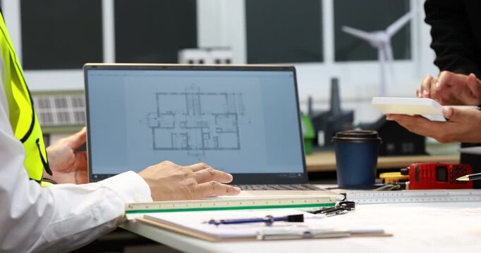 Engineers review a building floor plan on a laptop at a worktable with measuring tools, notebook, coffee and safety vest in a modern office.