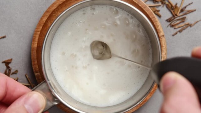 Hand froths oat milk in metal pitcher with electric whisk, creating foam for hojicha latte preparation on gray countertop.