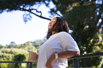 Adult woman leaning on balcony railing, wearing white top and smartwatch, holding mug, copy space © WavebreakMediaMicro