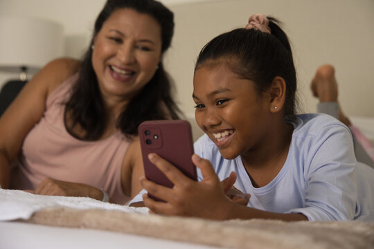 Mother and daughter reclining on bed, youth holding mauve smartphone and smiling warmly