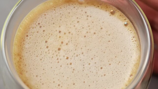 Hands slowly rotate glass of hojicha oat milk latte, revealing delicate creamy foam texture across surface before serving.
