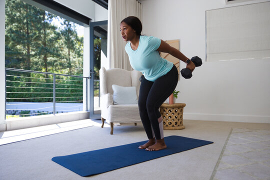 African woman performing bent-over dumbbell rows on blue mat in living room wearing fitness tracker