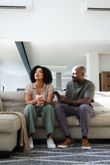 Fototapeta premium African American couple sitting on light gray sofa in open plan living area holding smartphone
