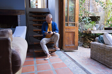Fototapeta premium Senior Asian man sitting on wooden stair in blue jacket mustard tee holding smartphone near patio