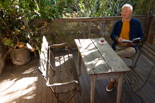 Asian senior sitting on deck right reading book with tea cup, wearing mustard shirt, copy space
