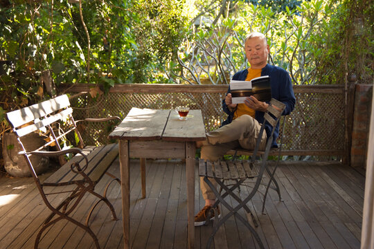 Senior Asian man sitting on porch reading book wearing mustard T-shirt tea on table, copy space
