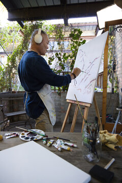 Senior Asian man standing at easel painting in garden studio, wearing stained apron and headphones