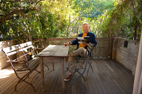 Senior Asian man reading book and holding glass at wooden table on terrace, wearing orange T-shirt