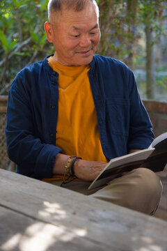 Senior Asian man sitting at wooden porch table reading book wearing mustard shirt and bracelets