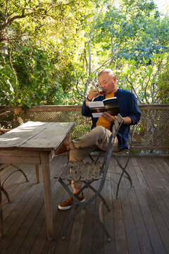 Senior Asian man sitting on folding chair on wooden deck reading book and sipping from cup