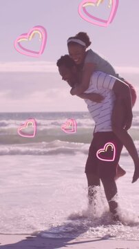 Animation of pink hearts over happy african american couple piggybacking on beach
