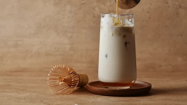 Hojicha pours over oat milk and ice in tall glass beside bamboo whisk, creating layered iced latte with syrup on brown set.