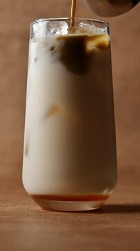 Hojicha tea pours over oat milk and ice in tall glass, creating layered iced latte during cold beverage preparation close up.