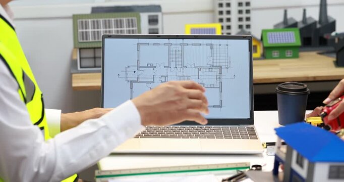 Architect reviewing a house floor plan on a laptop at a desk with model homes, drafting tools and a coffee cup.