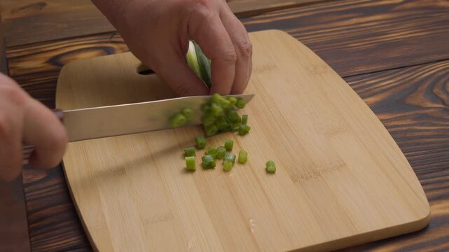 Close-up of a chef slicing fresh green onions with a kitchen knife on a wooden bamboo cutting board. Preparation of organic ingredients for cooking, culinary and food concept