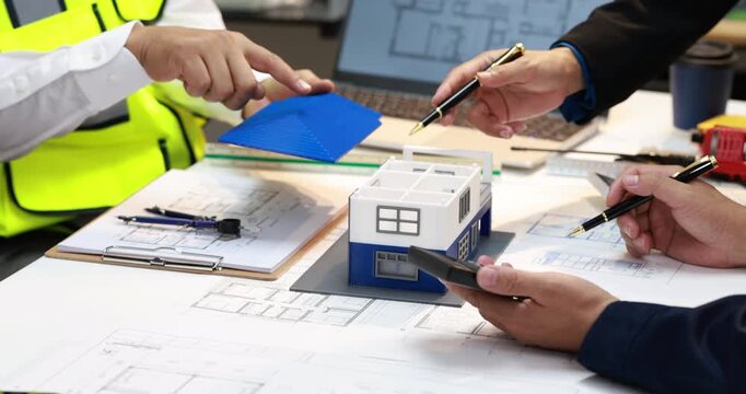 Architects and engineers review building plans around a table with a house model, blueprints, calculator, and safety vest during a construction planning meeting.
