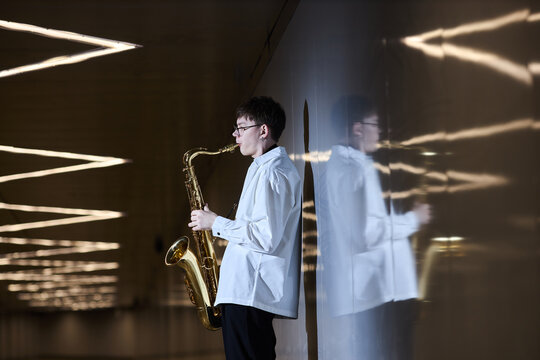 Young adult Caucasian man playing saxophone in underpass, standing near reflective wall. Image suitable for music school promotion, busking culture articles, or urban lifestyle campaigns