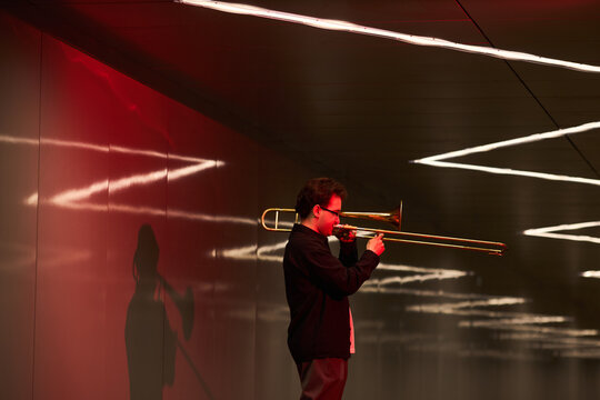 Young adult Caucasian man playing trombone in modern underpass, busking for urban entertainment. Image suitable for music education, street performance promotion, or cultural event marketing
