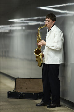 Vertical shot of young adult Caucasian man playing saxophone in subway station with open instrument case on floor, representing street performance and urban busking culture