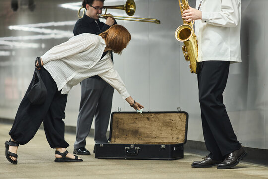 Caucasian woman putting money into instrument case of young adult male street musicians playing saxophone and trombone in subway. Concept of supporting local artists and busking culture