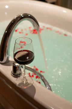 Glass of red wine by a romantic bathtub filled with water and rose petals.