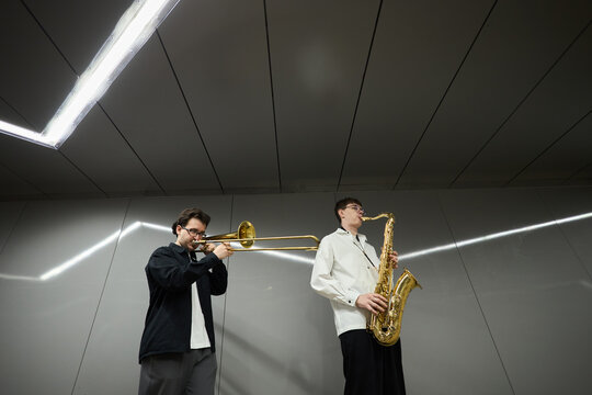 Two young adult Caucasian male street musicians playing trombone and saxophone in underground passage. Image suitable for music festival promotion or urban culture editorial, low angle shot