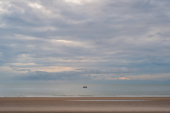 A lonely small fishing boat floating on the calm sea under cloudy sky and sandy beach.