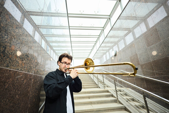 Young adult Caucasian man playing trombone in urban pedestrian underpass, busking for public. Image represents street performance culture and is suitable for music education marketing