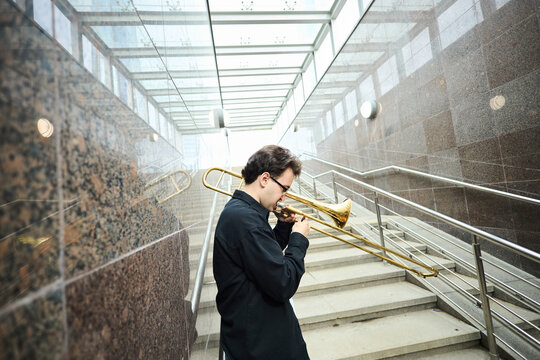 Young adult Caucasian man playing trombone on city stairs, performing as street musician. Image suitable for promoting urban culture, music education, and outdoor public events