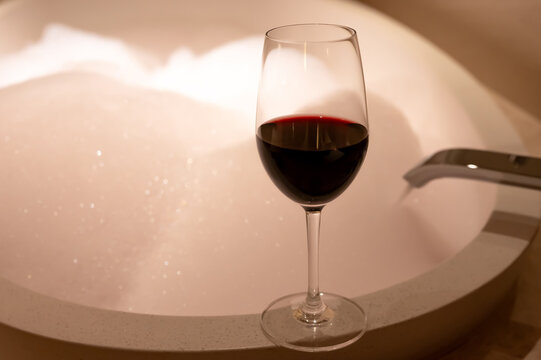 Glass of red wine on the edge of a round bathtub filled with bubble foam.