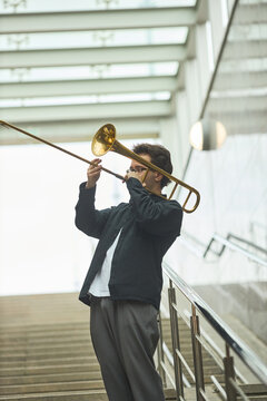 Vertical image of young adult Caucasian man playing trombone on city stairs. Image represents street performance and urban culture, suitable for music festival promotion