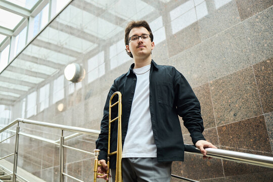 Portrait of young adult Caucasian male street musician holding trombone on urban stairs, looking at camera. Image suitable for music education and cultural event promotion, low angle view